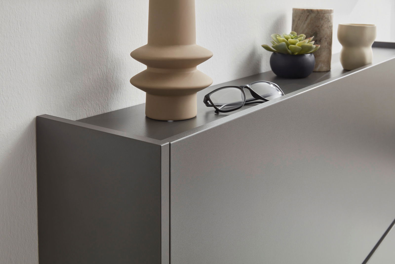 Nahaufnahme eines modernen, grauen Hängeschuhschranks mit Dekorationen wie einer Brille, einer Pflanze und Vasen auf der Oberseite, seitliche Perspektive.