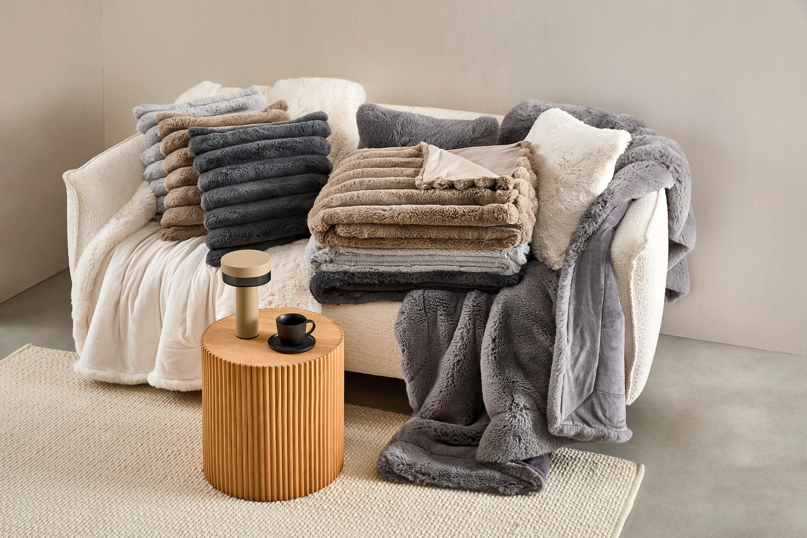 Ein gemütliches Wohnzimmer mit einer cremefarbenen Couch, die mit mehreren gestapelten Felldecken in verschiedenen Grautönen und Beige dekoriert ist. Auf einem kleinen Holztisch daneben steht eine Tasse und eine Lampe. Die Perspektive zeigt die Szene von 