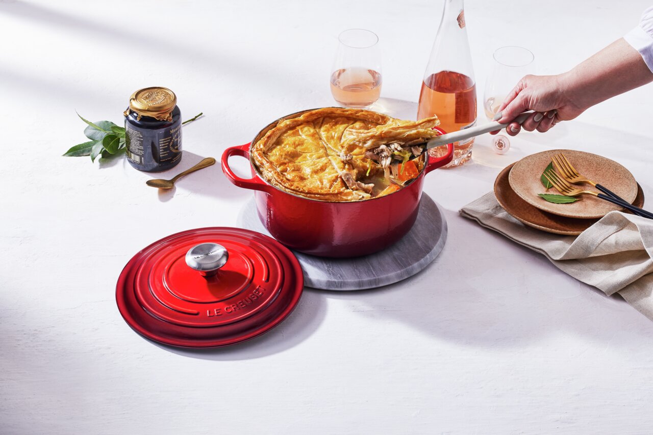 LE CREUSET Bräter Rund 26cm Kirschrot SIGNATURE Runder roter Bräter mit goldbrauner Kruste, von oben fotografiert, mit abgenommenem Deckel und einer Hand, die eine Portion serviert.