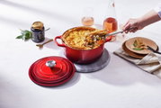 Runder roter Bräter mit goldbrauner Kruste, von oben fotografiert, mit abgenommenem Deckel und einer Hand, die eine Portion serviert.