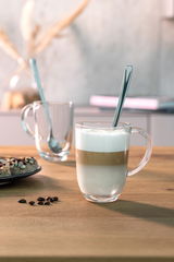 Latte Macchiato Tasse mit Löffel, gefüllt mit geschichtetem Kaffee und Milchschaum, auf einem Holztisch von vorne fotografiert.