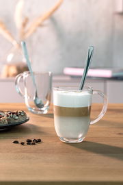 Latte Macchiato Tasse mit Löffel, gefüllt mit geschichtetem Kaffee und Milchschaum, auf einem Holztisch von vorne fotografiert.