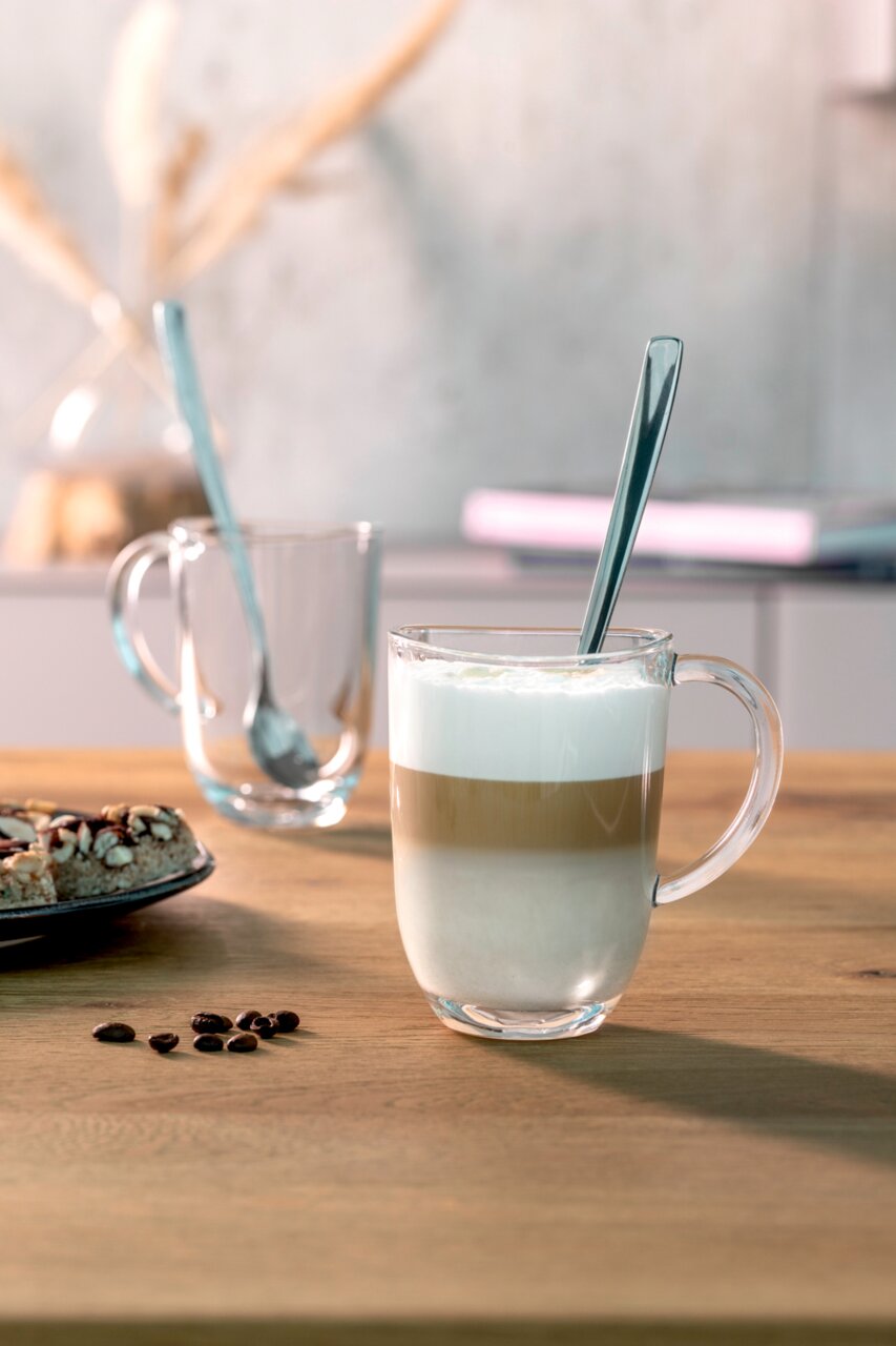 Latte Macchiato Tasse mit Löffel, gefüllt mit geschichtetem Kaffee und Milchschaum, auf einem Holztisch von vorne fotografiert.