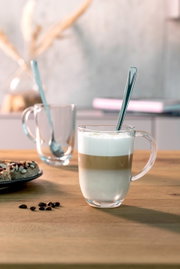 Latte Macchiato Tasse mit Löffel, gefüllt mit geschichtetem Kaffee und Milchschaum, auf einem Holztisch von vorne fotografiert.