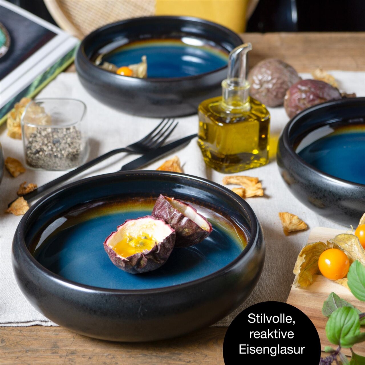 Vogelperspectief op een stijlvol, blauw-bruin soepbord met een reactief ijzerglazuur, gevuld met een doorgesneden passievrucht, omringd door andere borden, kruiden en decoraties op een tafel.