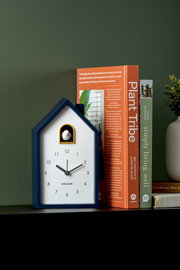 Moderner Wecker in Hausform mit Kuckuck, frontal auf einem Regal neben Büchern platziert.