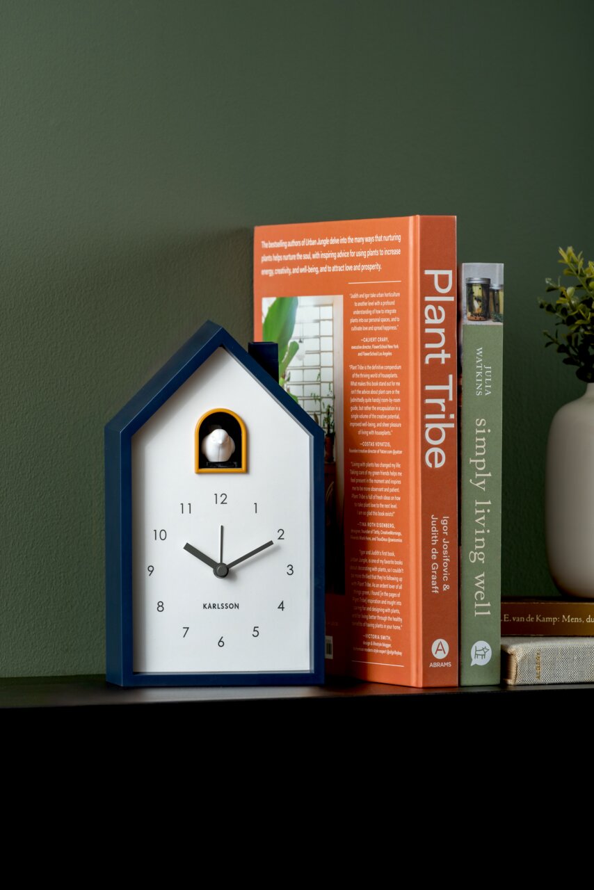 Moderner Wecker in Hausform mit Kuckuck, frontal auf einem Regal neben Büchern platziert.