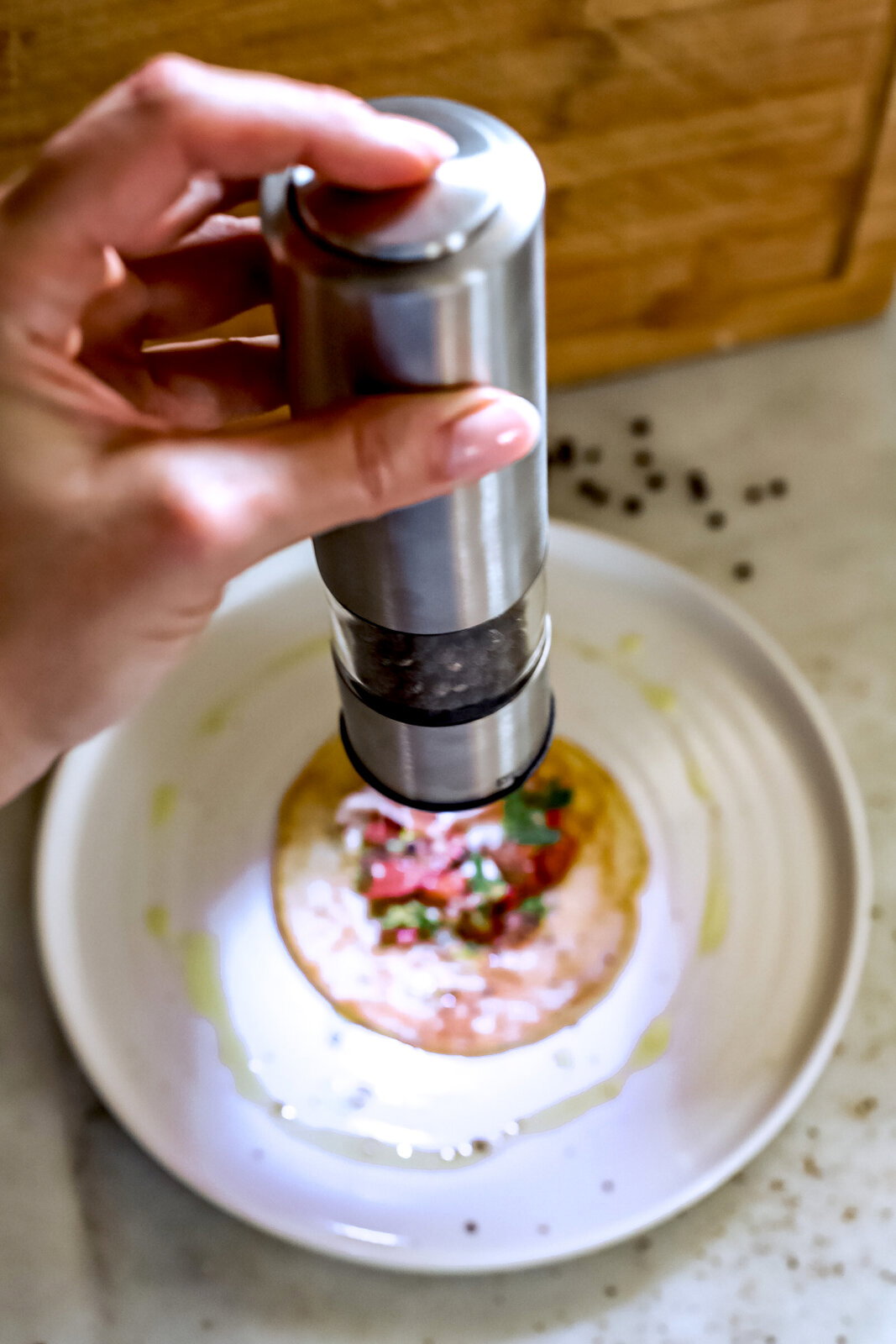 Nahaufnahme einer Hand, die eine silberne Pfeffer- oder Salzmühle über einem Teller mit Essen hält. Die Mühle wird von oben betrachtet.