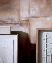 Frontaal detailaanzicht van minimalistische witte reliëfkunst in naturel houten lijsten, stijlvol geplaatst tegen een warme, rustiek gestucte muur.