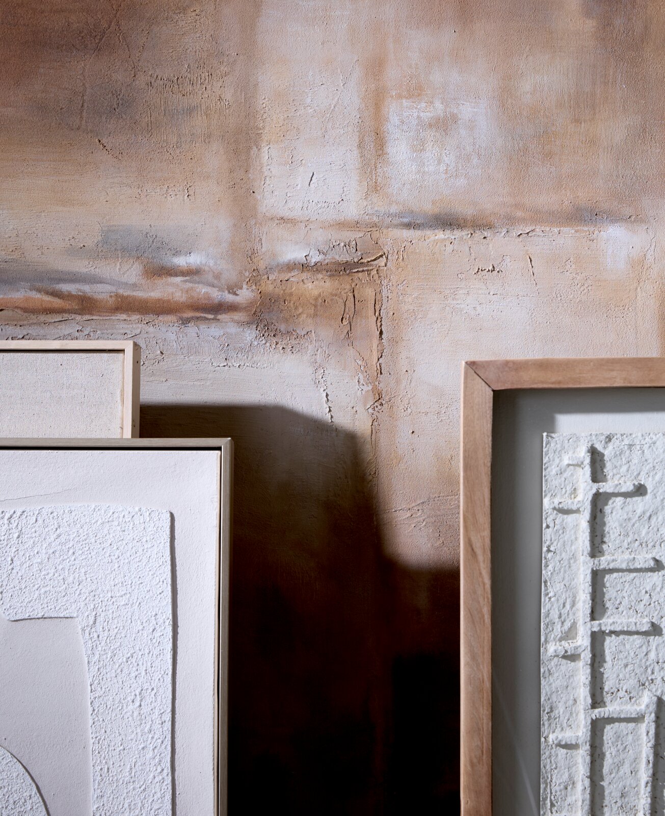 Frontaal detailaanzicht van minimalistische witte reliëfkunst in naturel houten lijsten, stijlvol geplaatst tegen een warme, rustiek gestucte muur.