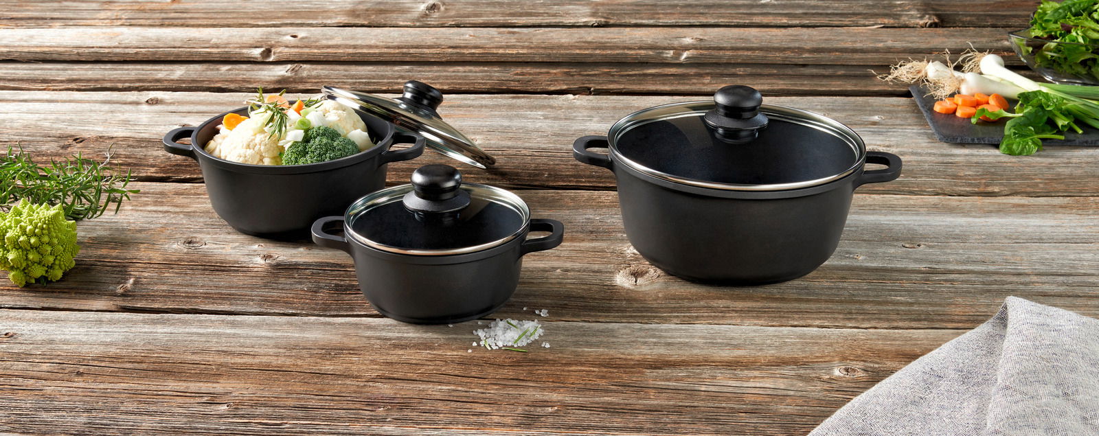 Dreiteiliges Topfset aus schwarzem Material mit Glasdeckeln, gefüllt mit frischem Gemüse, auf einem rustikalen Holztisch von oben betrachtet.