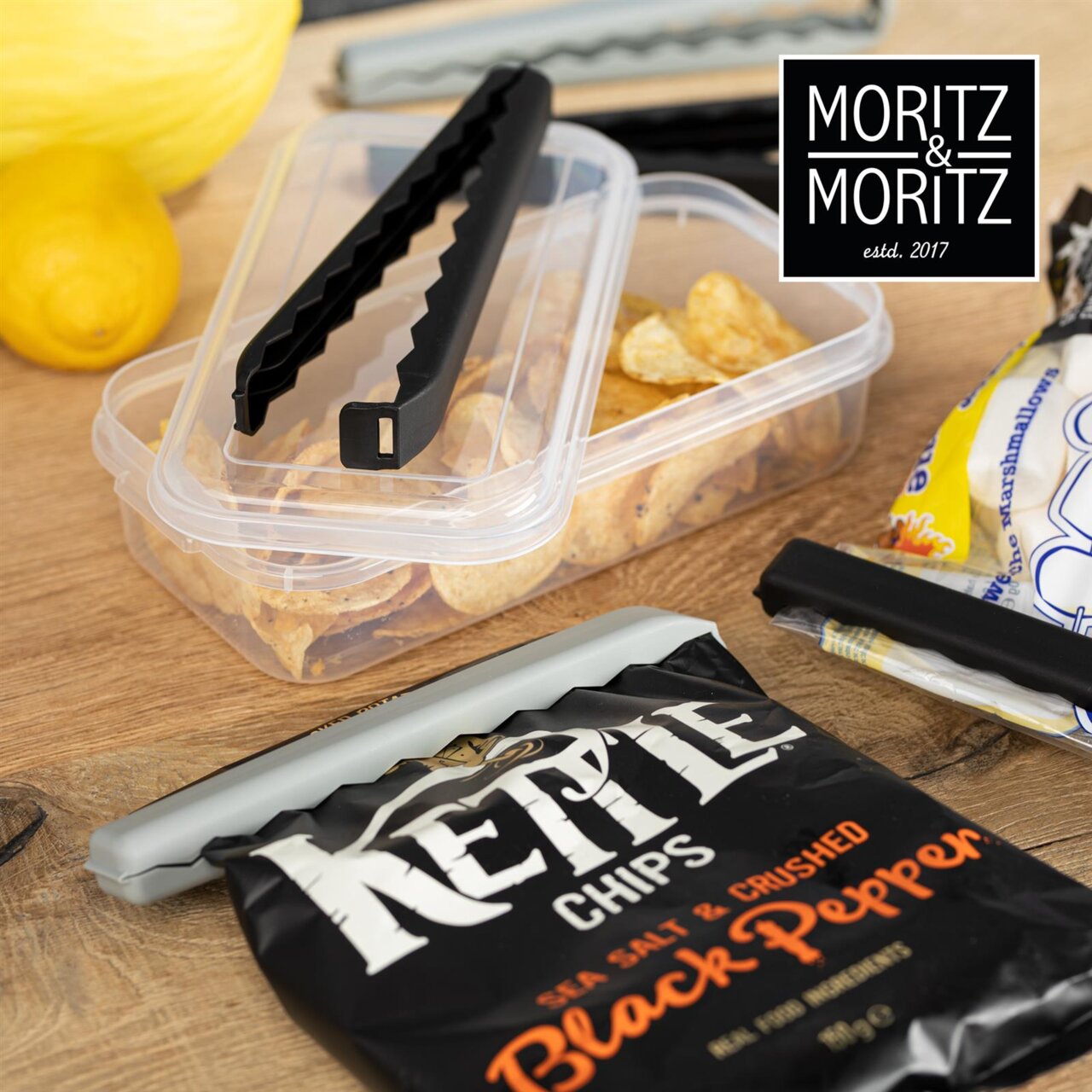 Verschlussclips in XXL-Größe, die auf einer geöffneten Chipstüte und einer Frischhaltebox mit Chips zu sehen sind. Perspektive von oben.