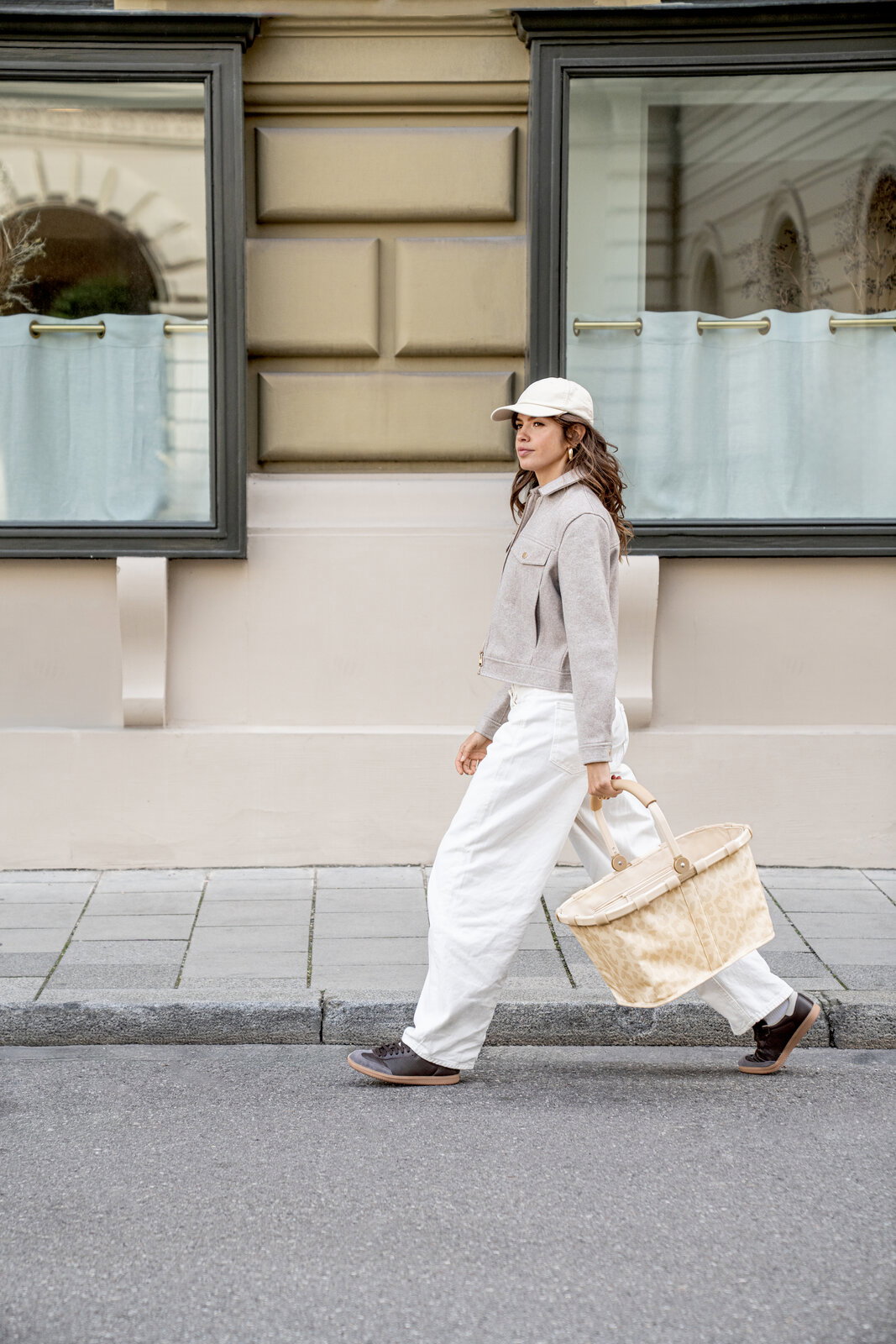 Seitenansicht: Frau geht auf dem Gehweg und trägt einen großen beigefarbenen Korb mit Muster und Tragegriffen vor einer Hausfassade mit Fenstern.