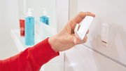 Nahaufnahme einer Hand, die einen weißen tesa Powerstrips Haken L Ecki an einer glatten Oberfläche anbringt, im Hintergrund sind Badezimmerutensilien zu sehen.