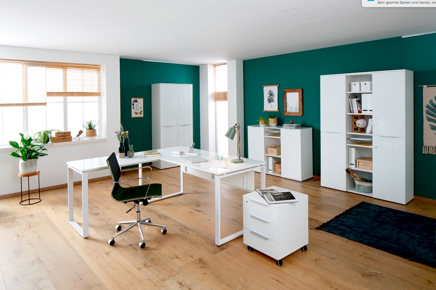 Moderne Büromöbelkombination in einem hellen Raum mit grün gestrichener Wand, bestehend aus einem weißen Schreibtisch, Rollcontainer, Aktenschränken und Sideboard. Perspektive: Frontalansicht.