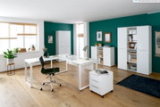 Moderne Büromöbelkombination in einem hellen Raum mit grün gestrichener Wand, bestehend aus einem weißen Schreibtisch, Rollcontainer, Aktenschränken und Sideboard. Perspektive: Frontalansicht.
