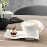 Weiße Kaffeetasse mit geschwungenem Henkel auf einem passenden Teller, gefüllt mit Café au Lait, von schräg oben fotografiert. Daneben ein kleines Dessert.