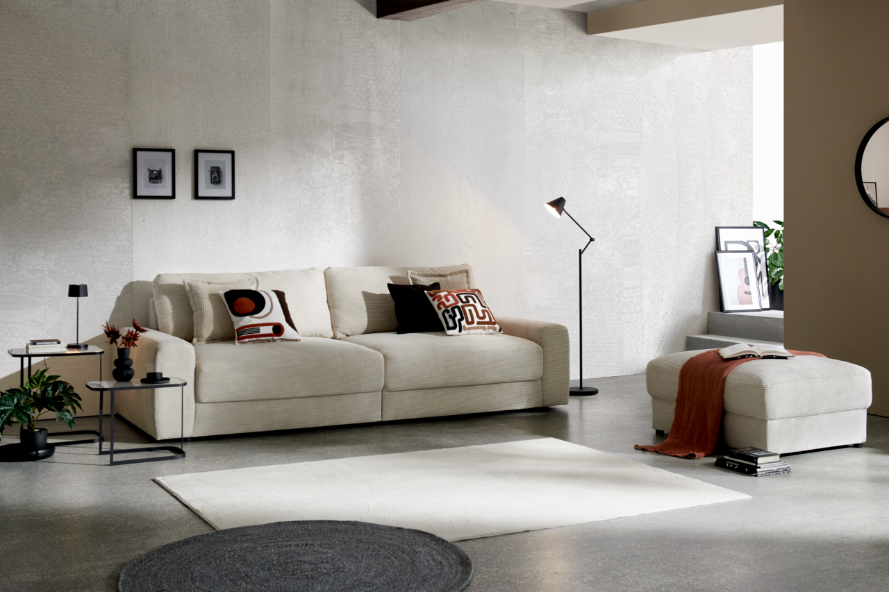 Großes, cremefarbenes Big-Sofa in modernem Wohnzimmer, seitliche Perspektive, mit dekorativen Kissen und passendem Hocker.