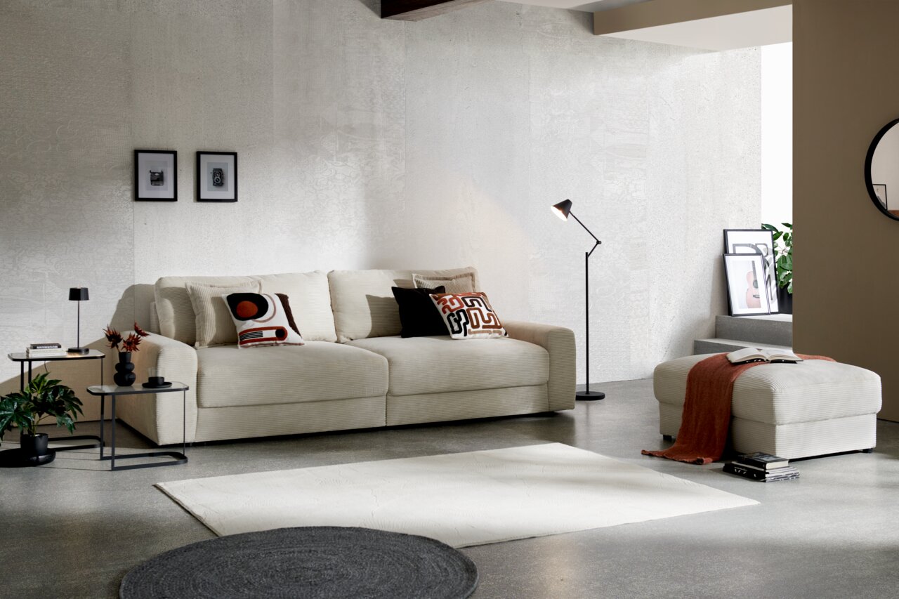 Z2 Big-Sofa Napoli Großes, cremefarbenes Big-Sofa in modernem Wohnzimmer, seitliche Perspektive, mit dekorativen Kissen und passendem Hocker.