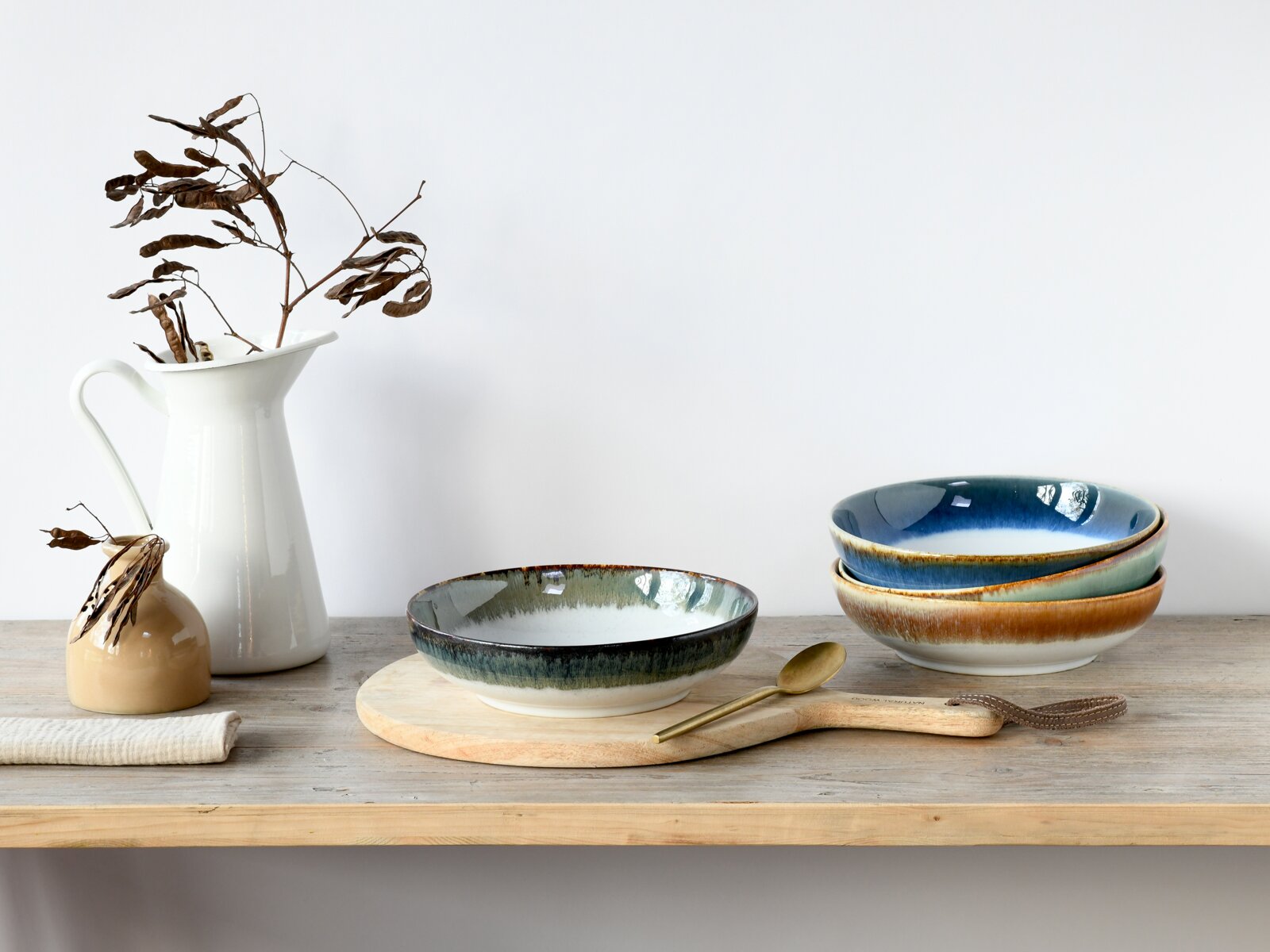 Keramikschalen mit blau-grünem Verlaufsglasur-Design auf Holzbrett und Holztisch, daneben Messinglöffel und weiße Kanne mit Trockenästen, Seitenansicht auf Augenhöhe