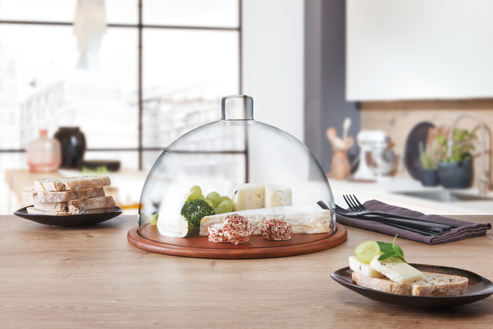 Holzkäseplatte mit Glasglocke, gefüllt mit verschiedenen Käsesorten und Trauben, auf einem Tisch in einer modernen Küche, seitliche Perspektive