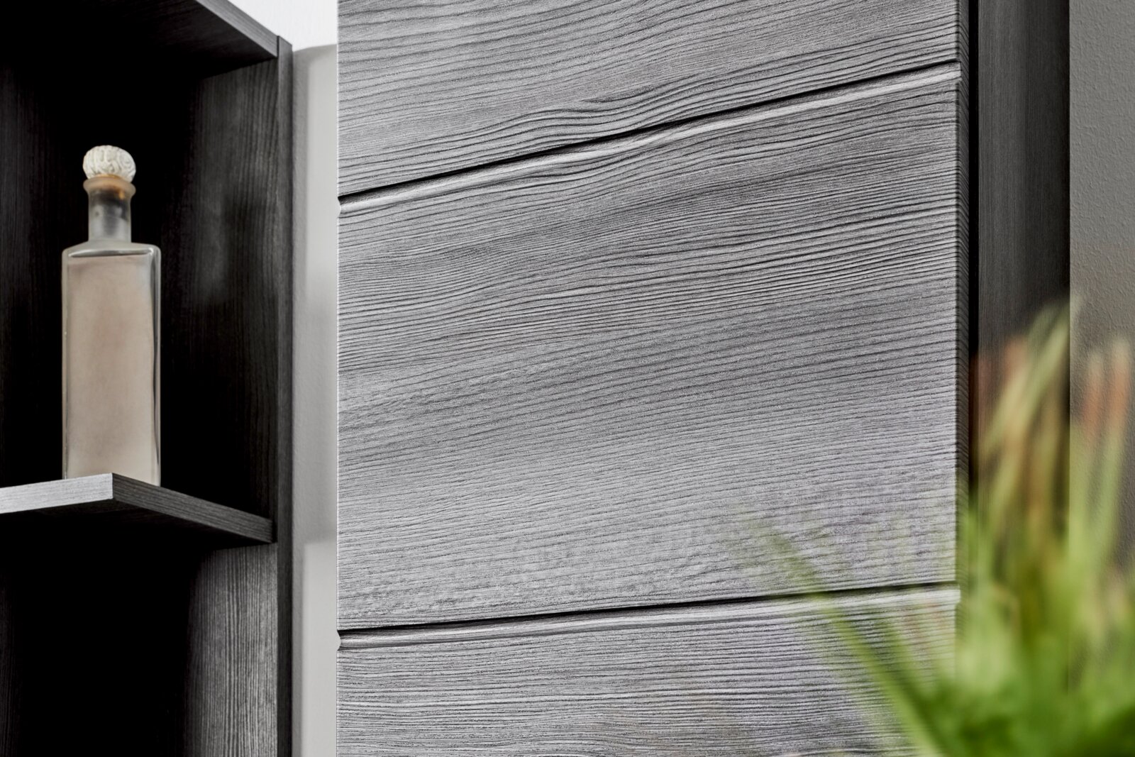 Detailaufnahme aus leicht seitlicher Frontperspektive: graue Holzmaserungs-Schrankfront mit horizontalen Fugen neben offenem Regal mit Glasflasche