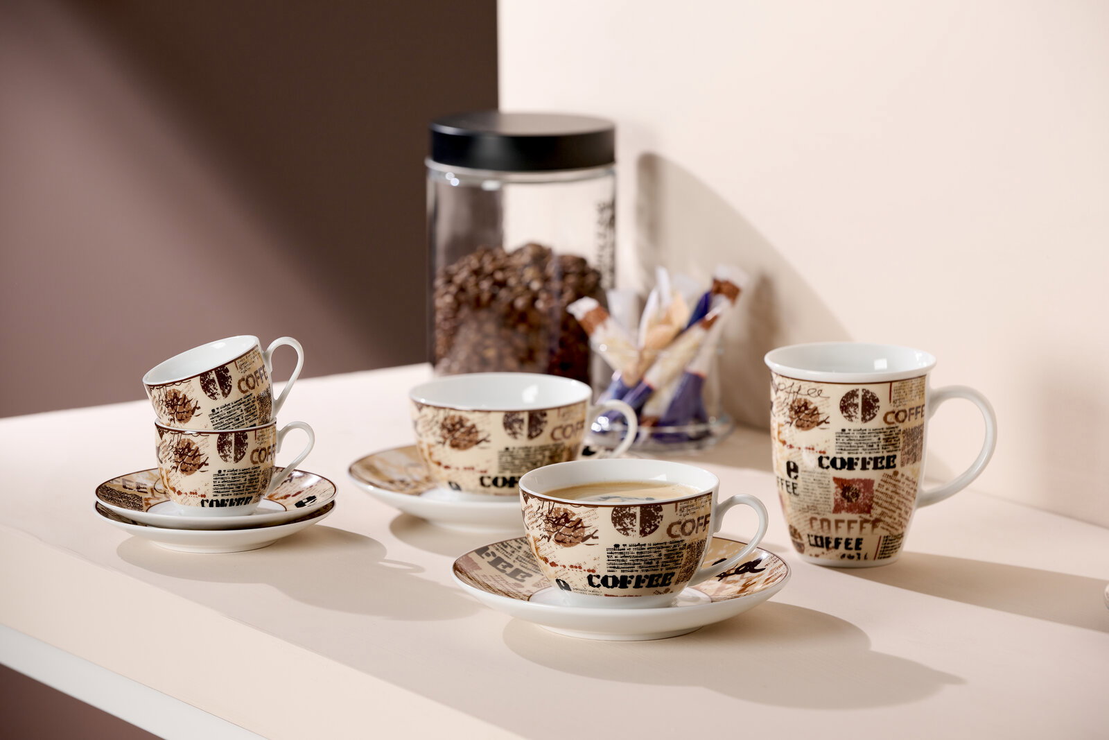 Ritzenhoff & Breker Kaffeebecher CREMA Vier Kaffeetassen mit Untertassen, dekoriert mit Kaffeebohnen- und Schriftmotiven, auf einem Tisch arrangiert. Im Hintergrund ein Glasbehälter mit Kaffeebohnen und Zuckersticks. Perspektive von schräg oben.