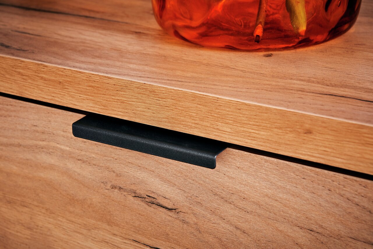 Close-up van een houten dressoir met een zwarte handgreep en een decoratieve glazen vaas in warme kleuren op het blad.