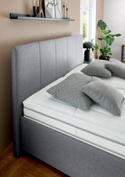 Grijs gestoffeerd bed met hoog hoofdeinde, gezien vanaf de zijkant, versierd met geometrische kussens en planten op de achtergrond.