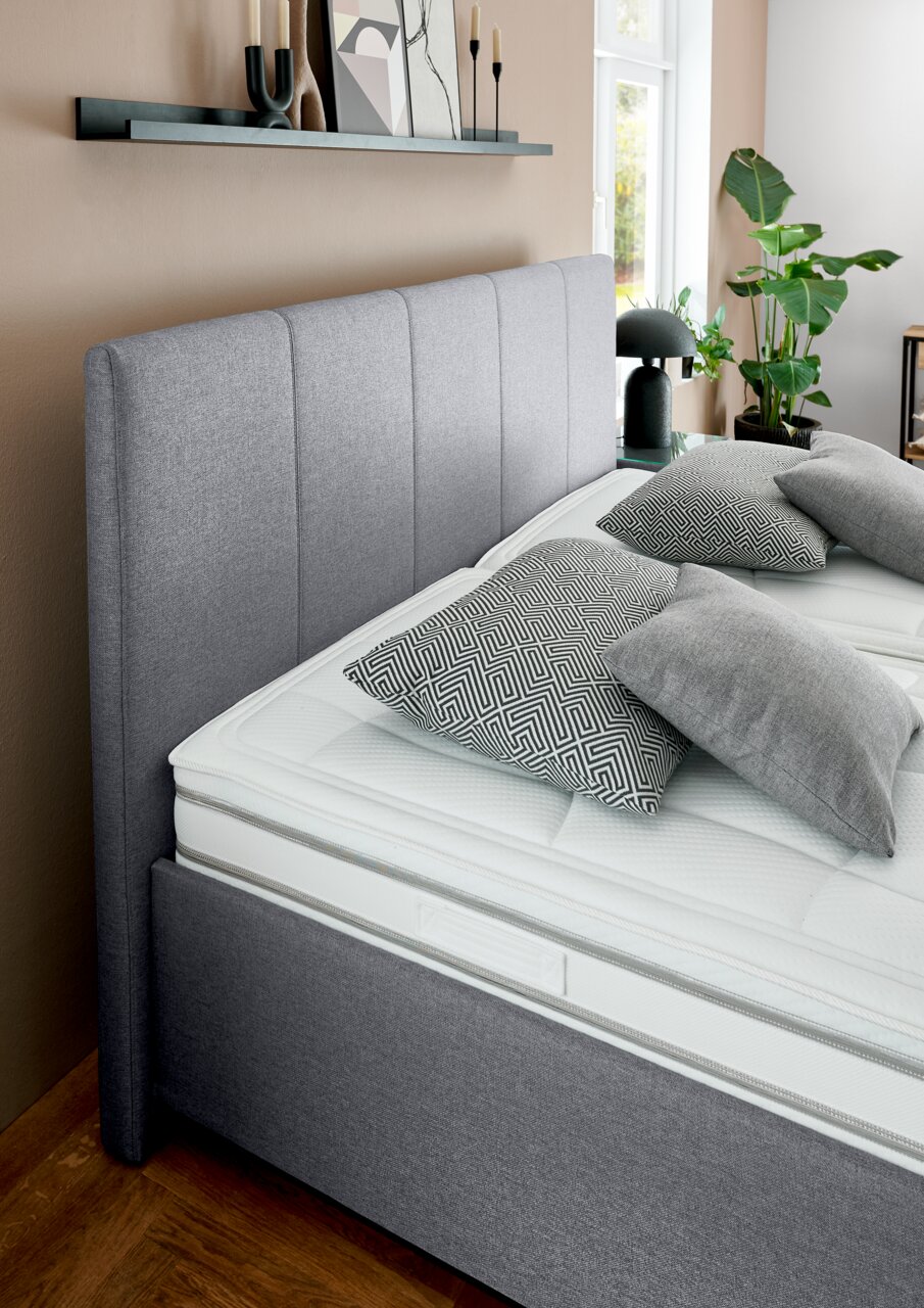 Grijs gestoffeerd bed met hoog hoofdeinde, gezien vanaf de zijkant, versierd met geometrische kussens en planten op de achtergrond.