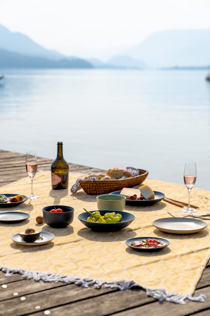 Thomas Dinerbord NATURE Ein gedeckter Tisch auf einem Steg mit Blick auf einen See, Teller und Schalen mit Obst und Käse, sommerliche Picknick-Atmosphäre.