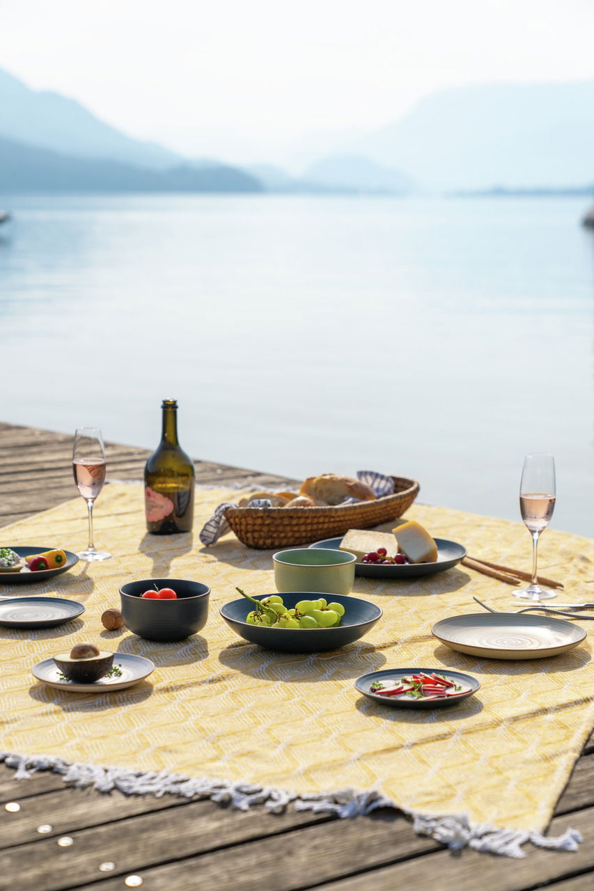 Thomas Speiseteller NATURE Ein gedeckter Tisch auf einem Steg mit Blick auf einen See, Teller und Schalen mit Obst und Käse, sommerliche Picknick-Atmosphäre.