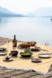 Ein gedeckter Tisch auf einem Steg mit Blick auf einen See, Teller und Schalen mit Obst und Käse, sommerliche Picknick-Atmosphäre.