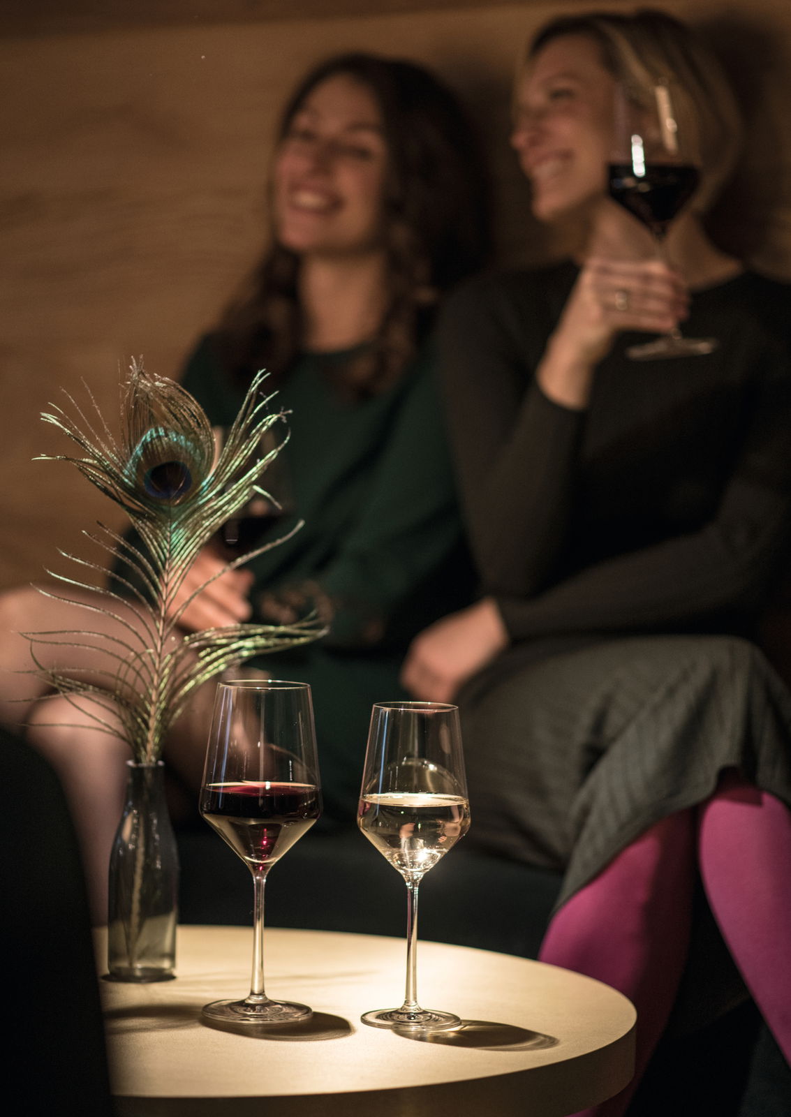 Zwei Sauvignongläser auf einem Tisch, gefüllt mit Rot- und Weißwein, im Vordergrund mit unscharfem Hintergrund von zwei Personen in entspannter Atmosphäre.