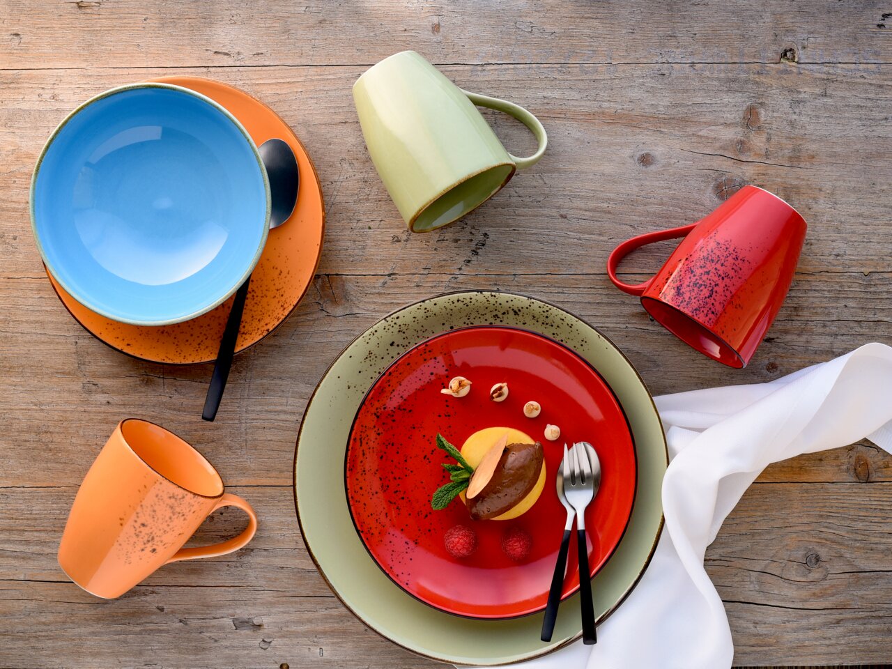 Bunte Geschirrkollektion aus der Vogelperspektive, bestehend aus einem blauen Teller auf einem orangefarbenen Teller, einer grünen Tasse, einer roten Tasse und einem roten Teller mit Dessert.