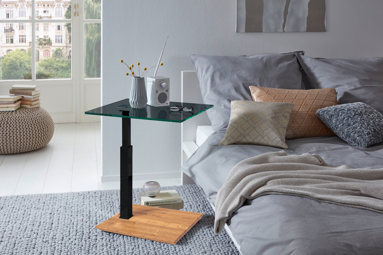 Moderner Beistelltisch mit Glasplatte und Holzfuß, seitliche Perspektive neben einem Bett mit Kissen und Decke.