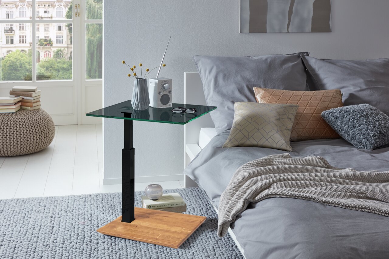 Moderner Beistelltisch mit Glasplatte und Holzfuß, seitliche Perspektive neben einem Bett mit Kissen und Decke.