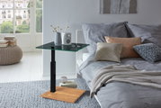 Moderner Beistelltisch mit Glasplatte und Holzfuß, seitliche Perspektive neben einem Bett mit Kissen und Decke.