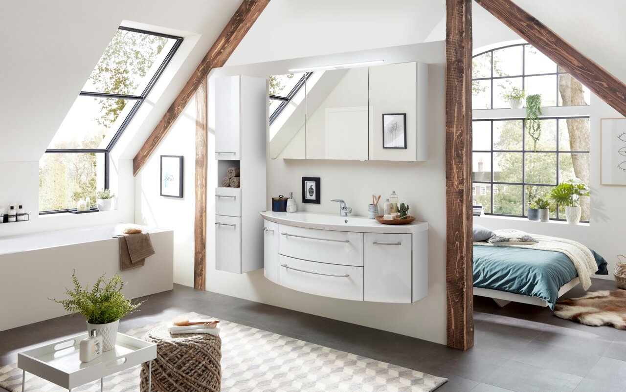 Sadena Badkombination 3-tlg. PLAKA Moderne Badkombination in einem hellen Dachgeschossbad mit großen Fenstern, bestehend aus einem weißen Waschtisch mit zwei Schubladen, einem hohen Schrank und einem Spiegelschrank. Perspektive von schräg vorne.