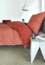 Rood satijnen beddengoed met een fijn patroon op een bed, gezien vanuit de zijkant, met nachtkastje en decoratie.
