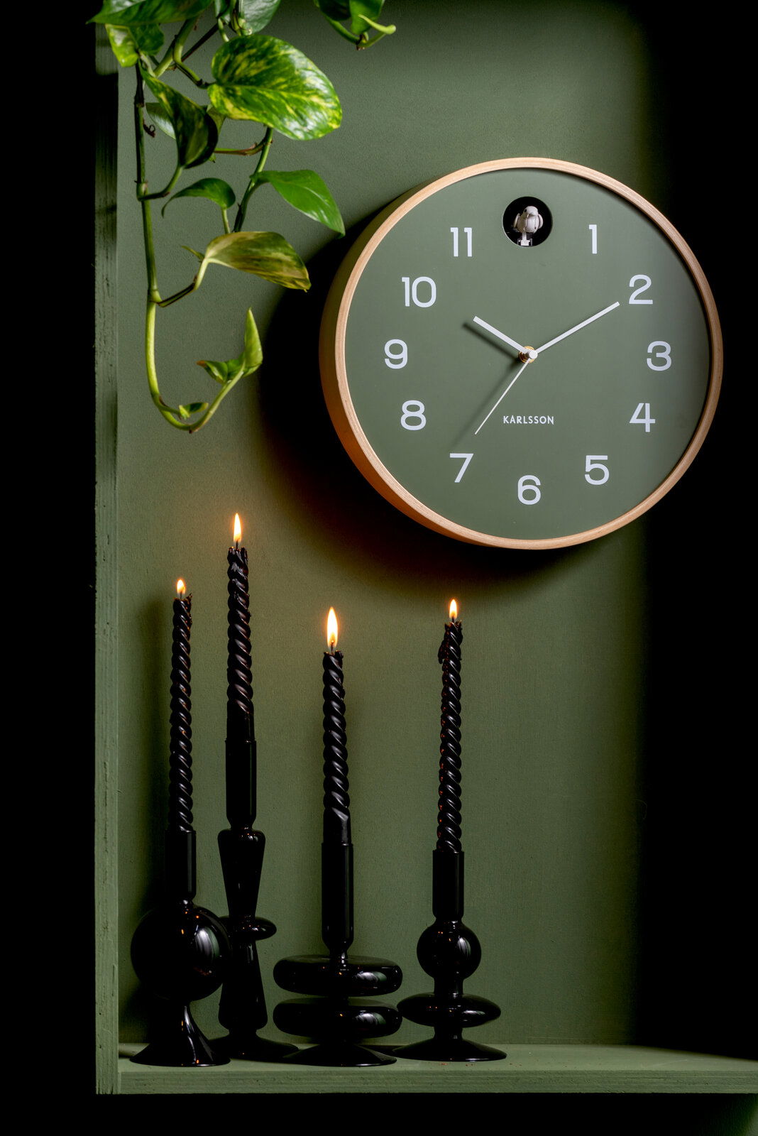 Grüne Wanduhr von KARLSSON mit Kuckuck, umgeben von Pflanzen und schwarzen Kerzen, aus der Frontalperspektive.