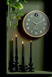 Grüne Wanduhr von KARLSSON mit Kuckuck, umgeben von Pflanzen und schwarzen Kerzen, aus der Frontalperspektive.