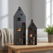Twee zwarte, huisvormige metalen lantaarns met raamopeningen en brandende theelichtjes, op een lichte houten tafel in de woonkamer, vooraanzicht licht schuin van voren.