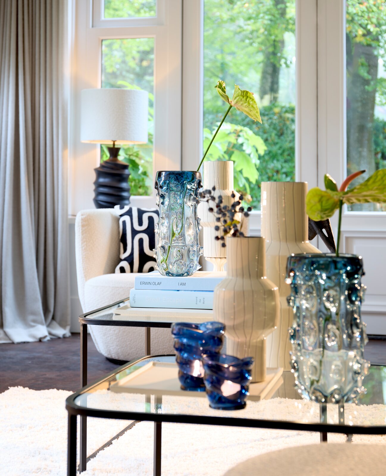 Dekorative Vasen aus strukturiertem blauem Glas und cremefarbener Keramik, aus leicht erhöhter frontaler Perspektive auf verspiegelten Couchtischen in einem hellen Wohnzimmer inszeniert.