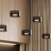 Moderne LED-Hängeleuchte mit schwarzem, zylindrischem Schirm, aus der Perspektive von unten betrachtet, vor einem Hintergrund aus Holz und Vorhängen.