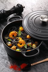 Schwarzer runder Bräter von Le Creuset mit geöffnetem Deckel, gefüllt mit schwarzer Pasta, Garnelen und Basilikumblättern, aus der Vogelperspektive fotografiert.