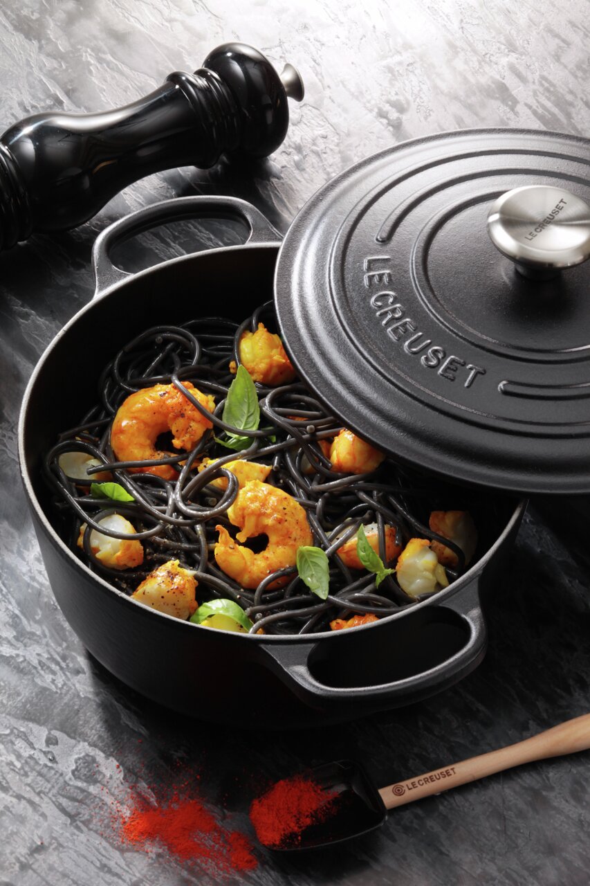 Schwarzer runder Bräter von Le Creuset mit geöffnetem Deckel, gefüllt mit schwarzer Pasta, Garnelen und Basilikumblättern, aus der Vogelperspektive fotografiert.
