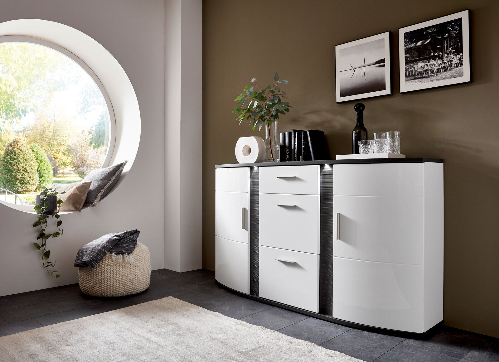Modernes weißes Sideboard mit geschwungenen Fronten, dekoriert mit Büchern, Pflanzen und Gläsern, in einem stilvoll eingerichteten Raum mit runder Fensteröffnung, seitliche Perspektive.