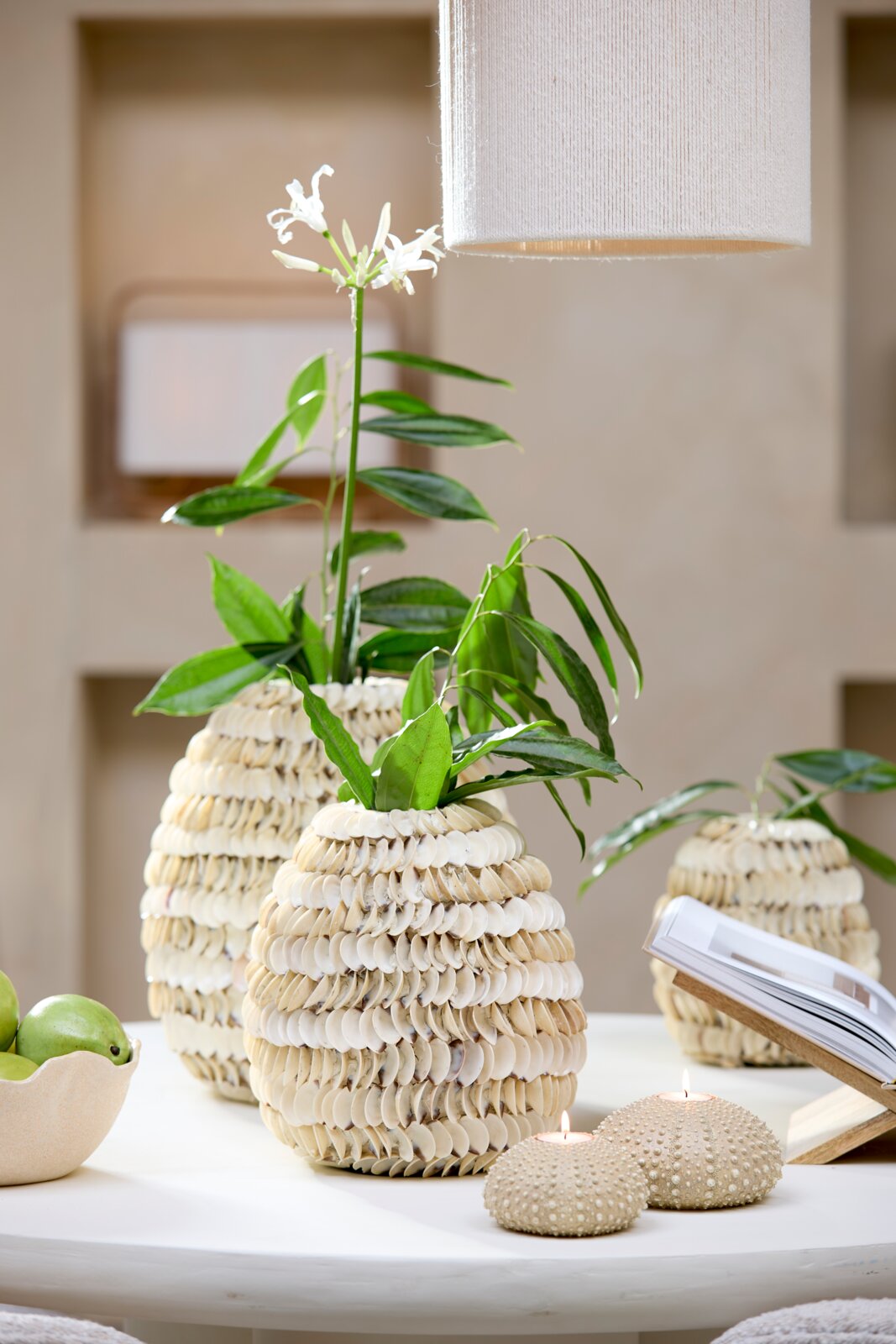 Vase mit Muscheldekor, gefüllt mit grünen Blättern und weißen Blüten, auf einem Tisch aus der Seitenperspektive.
