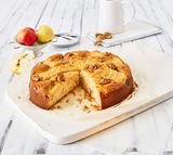 Ein Apfelkuchen mit Walnüssen auf einem weißen Holzbrett, von oben fotografiert. Im Hintergrund sind Äpfel, ein Messer und eine weiße Kanne zu sehen.
